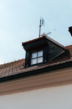 Geleneksel bir binanın eğimli çatısına tünemiş ahşap bir pencere, bir anten içerir ve gün ışığında açık mavi bir gökyüzü ile çevrilidir..