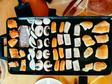 A grilling setup showcases assorted seafood and vegetables on a black griddle in a cozy dining atmosphere. Fresh ingredients are neatly organized for cooking. clipart