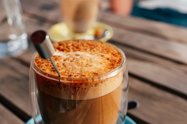 Köpükle kaplanmış zengin, kremalı kahve yumuşak, doğal ışık veren sakin bir kafe atmosferinde sade, ahşap bir masada, berrak bir cam bardakta oturur..