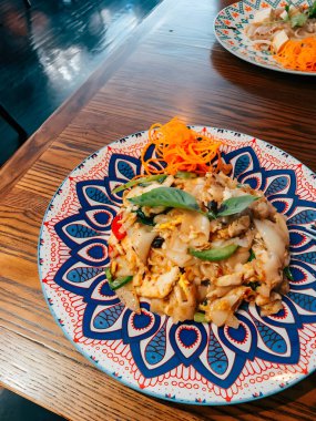 Taze tavuklu Tayland erişteleri, taze sebzeler ve aromatik otlar davetkar bir restoran atmosferinde dekoratif bir tabakta güzelce düzenlenmiştir..