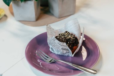 Hurma kremalı vejetaryen muzlu kek mor bir tabağa konur. Çörek kağıda sarılır ve yanında bir çatal ile birlikte gelir. Samimi bir ayar sunumu güçlendirir.