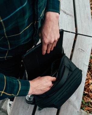 Bir insan, doğayla çevrili ahşap bir bankta otururken siyah bir sırt çantasından tablet alıyor. Odak noktası, açık havada gelişigüzel geçen bir anda cihazı geri alma eylemi..