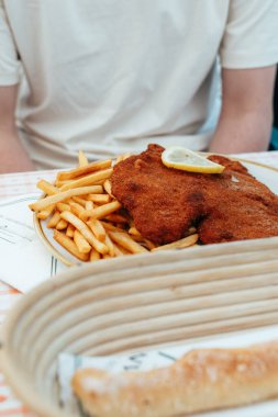 Limon dilimi ile süslenmiş lezzetli bir tabak çıtır şnitzel, cömert bir patates kızartması servisinin yanında duruyor. Sıradan bir atmosfer, bu klasik Avusturya yemeğinin tadını artırıyor..