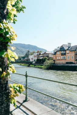 Bad Ischl 'deki huzurlu bir nehir kenarı, sahil boyunca doğa ile çevrili güzel binalar sergiliyor. Güneş ışığı suda parlıyor, bölgenin sakin atmosferini güçlendiriyor..