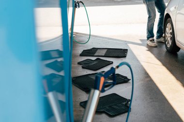 Bir kişi araba yıkama tesisinde paspasları hortumla temizliyor. Paspaslar durulanmak üzere yere serilirken, bir araç güneş ışığı altında yakınlarda park halindedir..