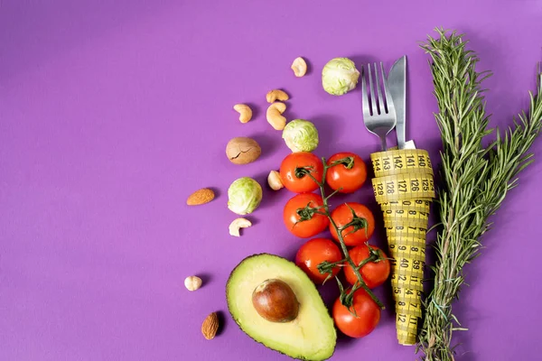 Avocados, cherry tomatoes, rosemary, nuts and cabbage lie on a purple background next to cutlery wrapped in measuring tape. Diet