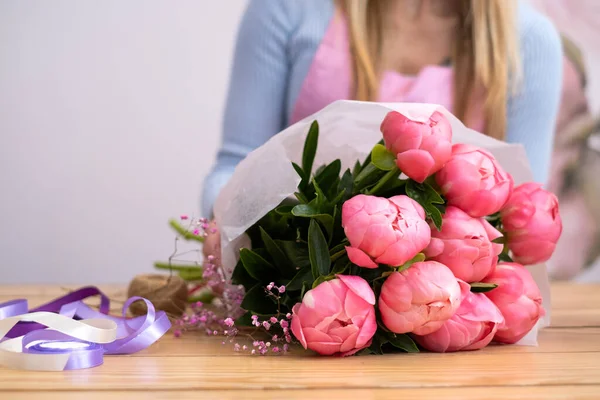 a girl florist holds a bouquet of pink peonies wrapped in white craft paper. Creation of beautiful bouquets. Flower Studio