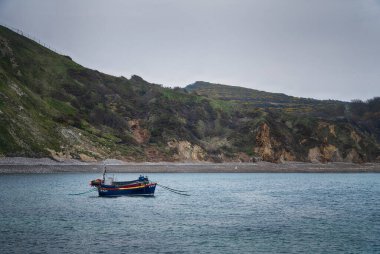İngiltere, Dorset - Nisan 2023: Bir balıkçı teknesi, Dorset 'teki Lulworth Koyu' nun sakin sularında, bulutlu bir gökyüzünün altında dramatik uçurumlarla dinleniyor..