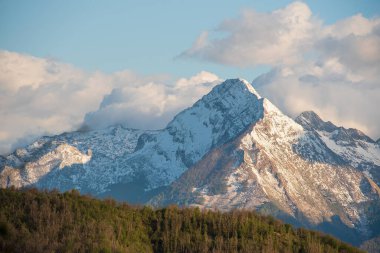 Emilia Romagna 'da Apennine Dağları' nın karlı zirveleri, dağılmış bulutlarla kaplı gökyüzünün altında, ormanlık tepelerin üzerinde yükselir..
