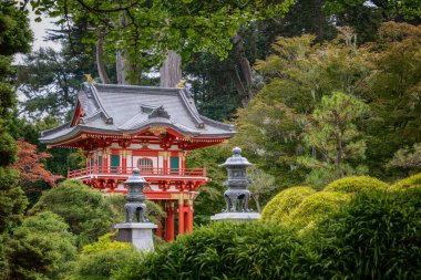 Huzurlu bir bahçede yeşil ve taş fenerlerle çevrili canlı kırmızı bir pagoda duruyor.