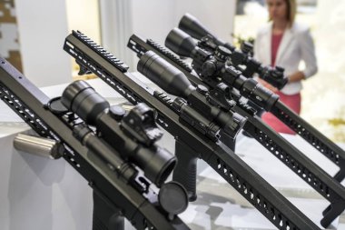 Silah fuarında sergilenen metal çubuğa monte edilmiş siyah dürbün optikleri, ayarlama düğmelerine yakın çekim detayları.