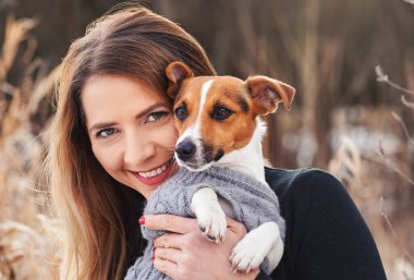 Genç bir kadın elinde Jack Russell teriyer köpeğini tutuyor. Elinde kışlık elbiseler var.