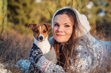 Kışlık ceketli genç bir kadın, karla kaplı zemine uzanmış, elinde teriyer köpeği, bulanık ağaçlar...