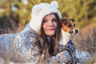 Kışlık ceketli genç bir kadın, karla kaplı zemine uzanmış, elinde teriyer köpeği, bulanık ağaçlar...