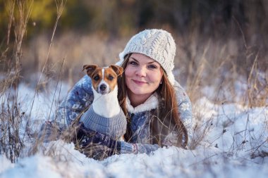 Kışlık ceketli genç bir kadın, karla kaplı zemine uzanmış, Jack Russell teriyer köpeğini tutuyor, bulanık ağaçlar arka planda