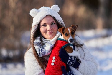 Kışlık ceketli genç bir kadın elinde Jack Russell teriyeri ile arka planda bulanık ağaçlar tutuyor.