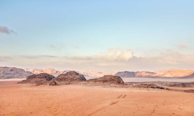 Turuncu kırmızı kum çölü, kayalık oluşumlar ve dağlar arka planda mavi gökyüzü, uzaktan görülebilen kamp çadırları - tipik Wadi Rum, Ürdün manzarası.