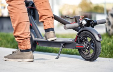 Park halindeki elektrikli scooter 'da insan ayağı, yakın çekim detayı, arka planda bulanık çimenler.