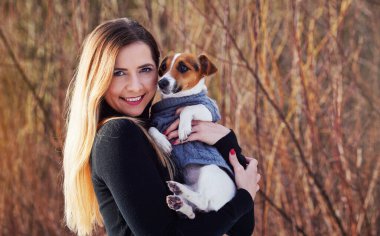 Genç bir kadın elinde Jack Russell teriyer köpeğini tutuyor. Elinde kışlık elbiseler var.
