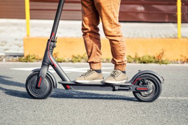 Ayaklarında elektrikli scooter ile asfalt yolda gezen sıradan pantolonlu genç bir adam.