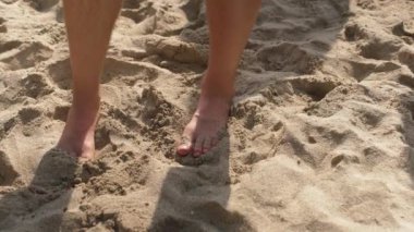 Genç adam kumlu sahilde dikiliyor, birkaç adım atıyor - ayaklarına yakın çekim ayrıntıları.