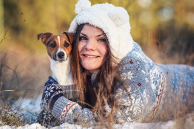 Kışlık ceketli genç bir kadın, karla kaplı zemine uzanmış, elinde teriyer köpeği, bulanık ağaçlar...