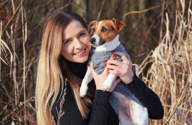 Genç bir kadın elinde Jack Russell teriyer köpeğini tutuyor. Elinde kışlık elbiseler var.
