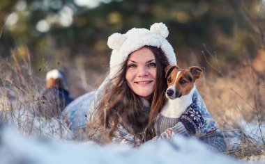 Kışlık ceketli genç bir kadın, karla kaplı zemine uzanmış, elinde teriyer köpeği, bulanık ağaçlar...