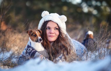Kışlık ceketli genç bir kadın, karla kaplı zemine uzanmış, elinde teriyer köpeği, bulanık ağaçlar...