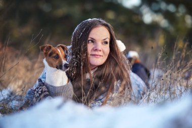 Kışlık ceketli genç bir kadın, karla kaplı zemine uzanmış, Jack Russell teriyer köpeğini tutuyor, bulanık ağaçlar arka planda