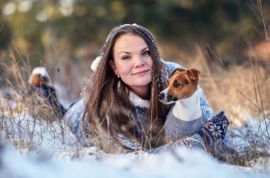 Kışlık ceketli genç bir kadın, karla kaplı zemine uzanmış, elinde teriyer köpeği, bulanık ağaçlar...