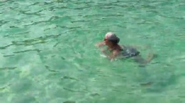 Gri saçlı yaşlı bir kadın, durgun yeşil denizde yüzüyor, yukarıdan manzaralı.