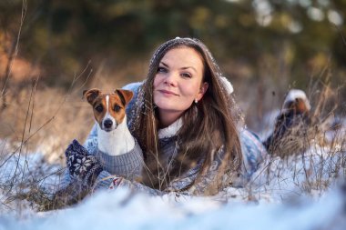 Kışlık ceketli genç bir kadın, karla kaplı zemine uzanmış, elinde teriyer köpeği, bulanık ağaçlar...