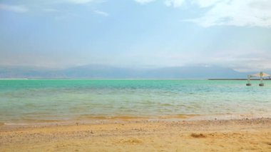 Ein Bokek 'te sakin bir gün, Ölü Deniz plajı, mavi yeşil su, yakınlardaki güneş gölgesi barınağı, kumsal ön planında güneş parlıyor.