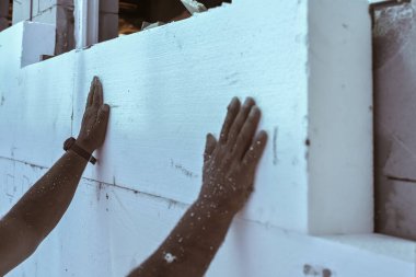 Çıplak ev duvarlarına polistiren ısı yalıtımı yükleyen işçiler, beyaz köpük tahtalarına elle dokunmak için detaylar.