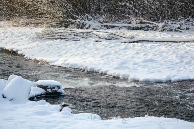 Kışın nehir, karlar kıyıların her iki tarafında, güneş arka planda ağaçların üzerinde parlıyor.