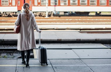 Uzun paltolu, tekerlekli siyah valizli genç bir kadın tren istasyonunda bekliyor.