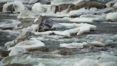 Kış nehri kar ve buzla kaplı taşların yakınından akıyor.