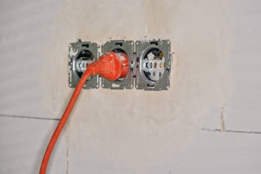 Ev inşaatı sırasında duvardaki geçici elektrik prizine bağlı turuncu kablo, yakın çekim detayı.