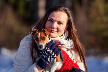 Kışlık ceketli genç bir kadın elinde Jack Russell teriyeri ile arka planda bulanık ağaçlar tutuyor.