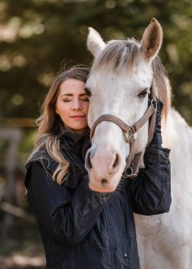 Beyaz Arap atına yaslanmış genç bir kadın, gözleri kapalı, arka planda bulanık ağaçlar, yakın plan ayrıntıları.