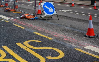 Mavi ok yön işareti, turuncu yol konilerinin yanında, asfalt yakınında sarı otobüs metni.