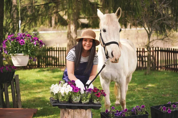 Woman in work clothes, gloves and straw hat working with flowers in garden, white horse near, blurred green trees and yard background