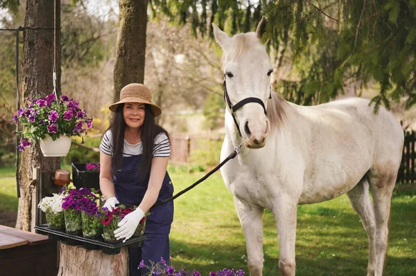 Woman in work clothes, gloves and straw hat working with flowers in garden, white Arabian horse near, blurred green trees and yard background