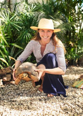 Young woman in straw hat and skirt, smiling - holding tortoise above ground in sun lit garden yard