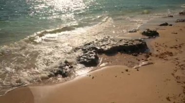 Güneşli bir günde kumsalda deniz suyuyla yıkanan küçük düzensiz taşlar, yakın çekim detayları.