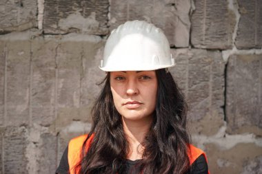 Beyaz kasketli, turuncu, yüksek görünürlüklü yelek giyen, uzun siyah saçlı, kameraya bakan genç bir kadın. Bulanık eski inşaat alanı arka planı