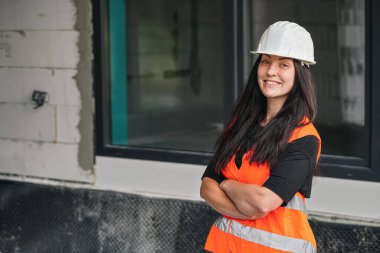 Beyaz kasketli, turuncu, yüksek görünürlüklü, uzun siyah saçlı, kameraya bakan, elleri çapraz güvenlikli genç bir kadın. Geniş pencere arkaplanlı bulanık inşaat alanı duvarı