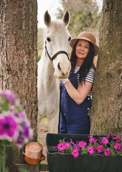Woman in working clothes and straw hat standing next to her white Arabian horse, trees on sides, blurred flowers foreground