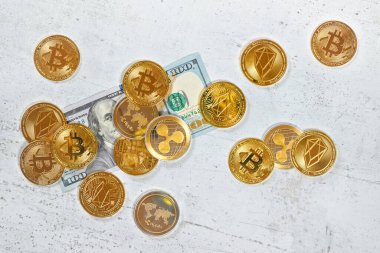 Üzerinde altın renkli kripto paralar olan 100 dolarlık banknot, yukarıdan görünen masa gibi beyaz taşlara saçılmış.